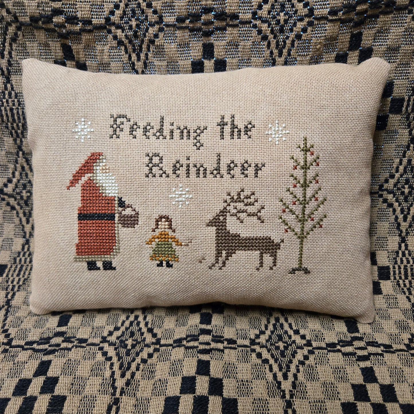 A square beige cross-stitch pillow featuring the text 'Feeling the Reindeer' and embroidered holiday figures including Santa, a reindeer, a girl, and a tree, displayed on a patterned couch.