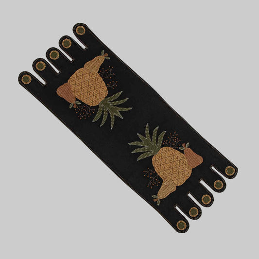 A black table runner with a folk art design featuring pineapples and tassels.