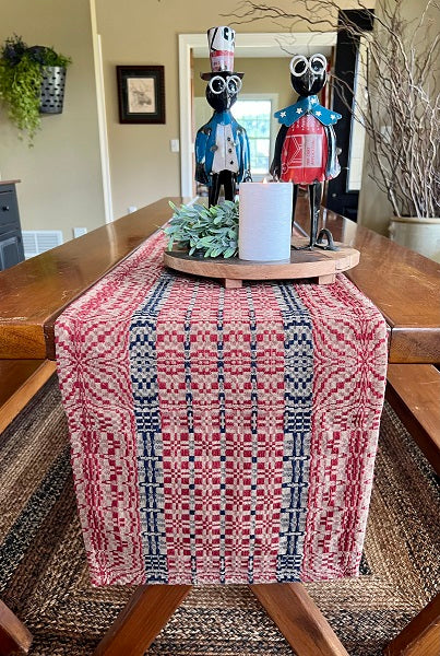 A short table runner with a red, blue, and tan geometric pattern, displayed on a wooden table with a decorative piece and a vase with flowers on top.