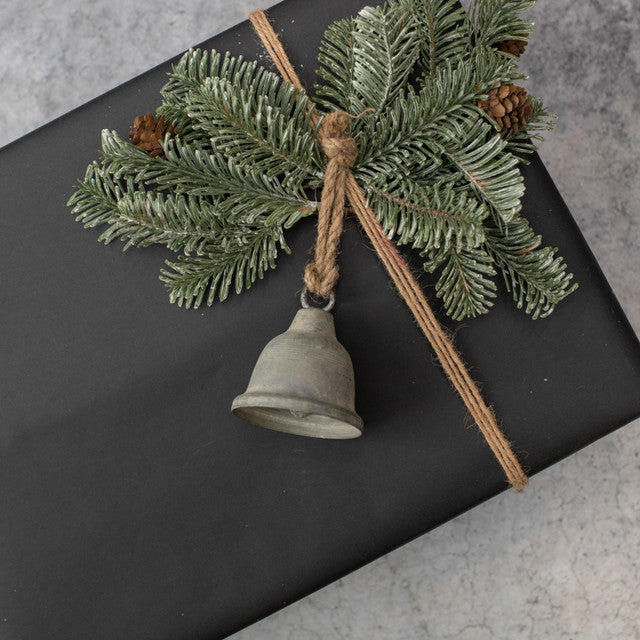 A galvanized metal bell ornament with a green pine branch and brown twine, placed on a black textured surface.