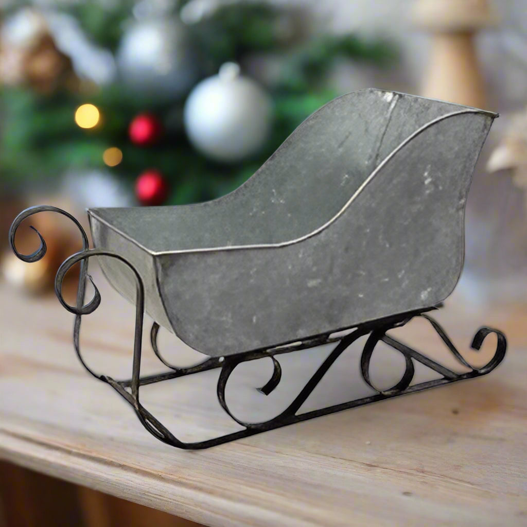 A silver galvanized metal sleigh with black metal runners and decorative swirled design at the bottom, displayed against a white background.