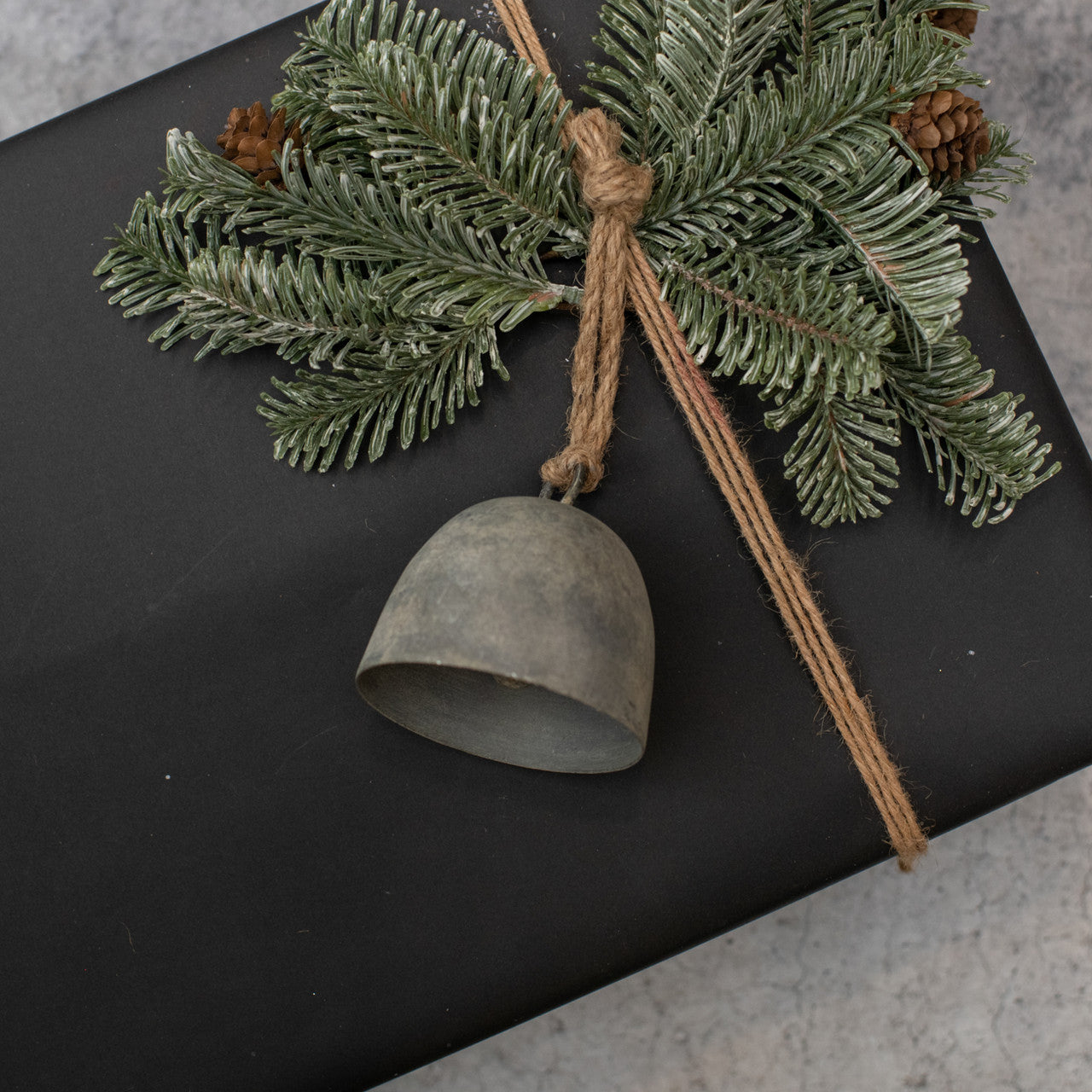 A silver galvanized Morgan bell ornament with a rustic finish, decorated with a pine branch and attached to a rope, placed on a black surface.