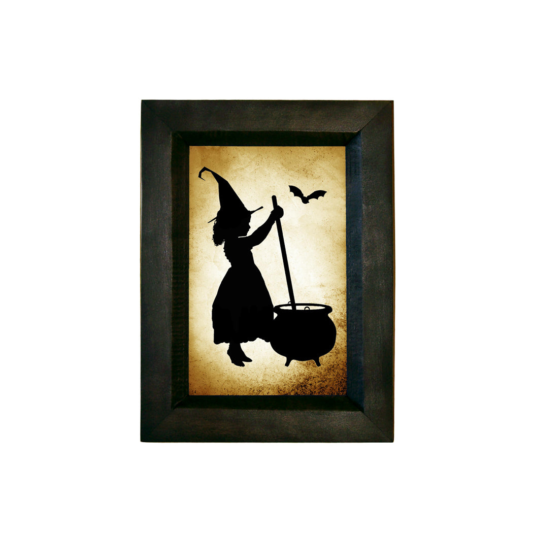 A silhouette of a girl witch stirring a pot, printed on high-quality paper with an antique appearance, framed in a black, solid distressed wood frame.