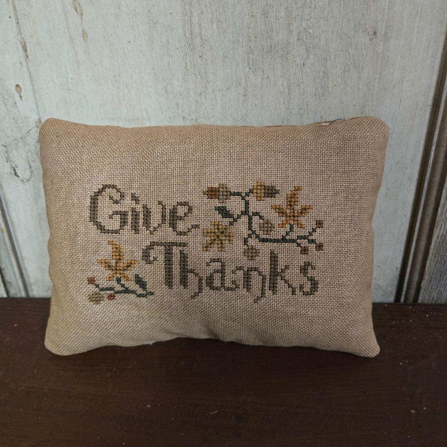 Small embroidered pillow with 'Give Thanks' text on a wooden surface