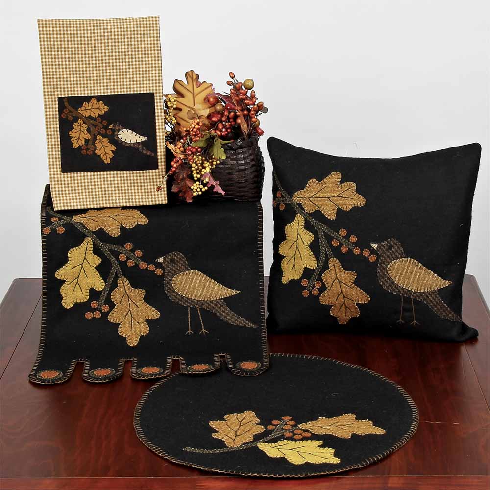 A set of table runners and pillows with a bird and leaf design, primarily in black, mustard, and golden colors, displayed on a wooden table.