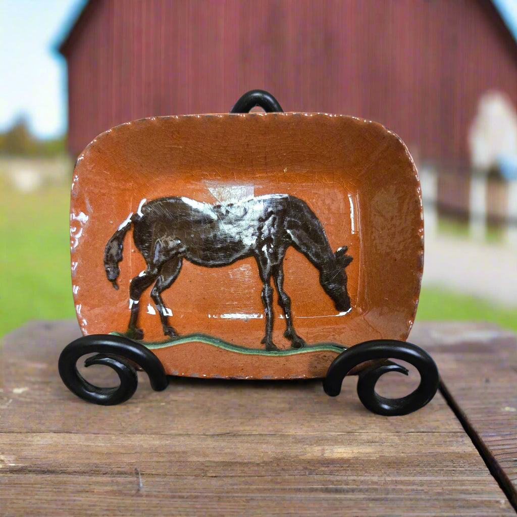A rectangular ceramic plate with a brown horse, displayed on a wooden surface with a barn in the background.