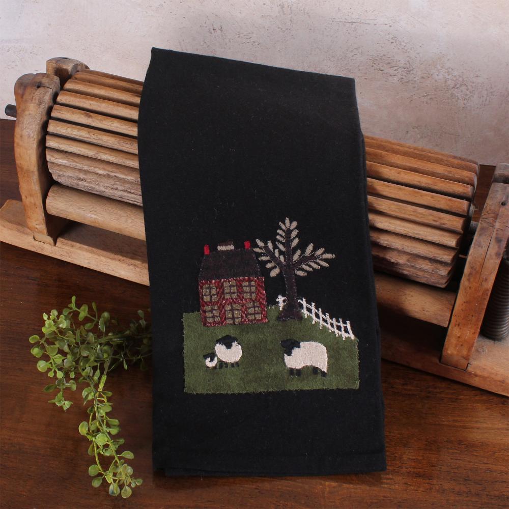 Black towel with embroidered design of a house, tree, and sheep on a wooden surface.