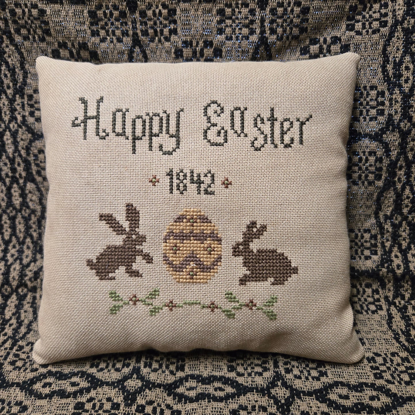 Decorative pillow with cross-stitched design of a rabbit and text 'Happy Easter 1842' on a textured fabric background.