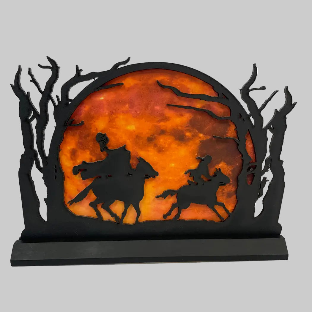 Decorative wall art with silhouettes of people on horseback against an orange moonlit sky, framed by black trees.