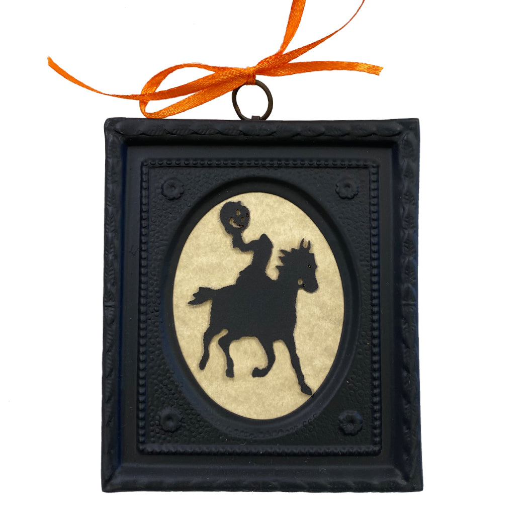 Decorative ornament with silhouette of a person on a horse in a black frame, hanging by an orange ribbon.