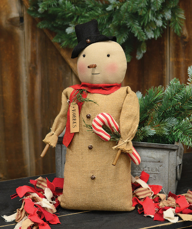 A plush toy resembling a snowman with a black top hat, red scarf, and brown paper tag that reads 'Hobbes'. It is decorated with jingle bells and has wooden details.