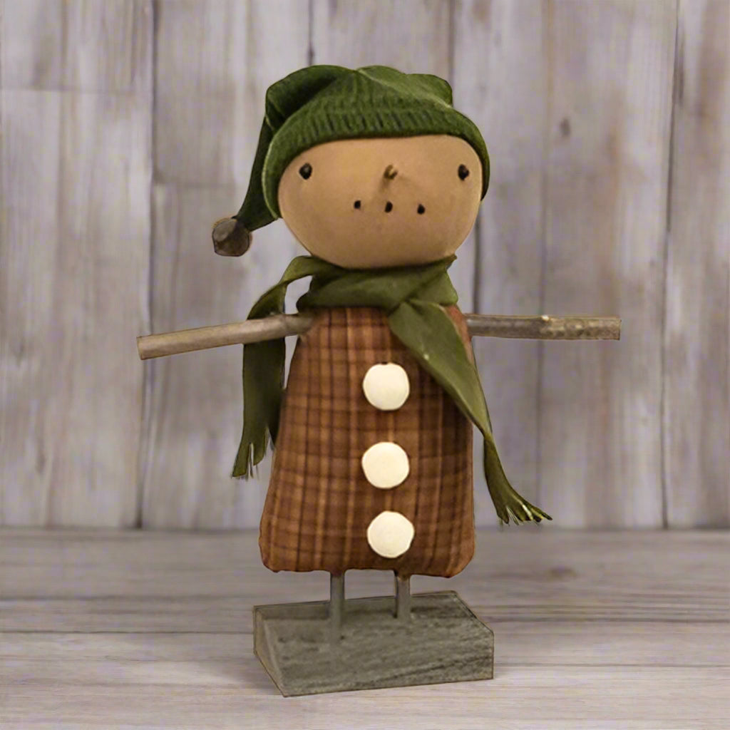 A plush doll resembling a snowman with a brown plaid dress, white pom buttons, a green scarf, and a winter hat, set on a wooden base.