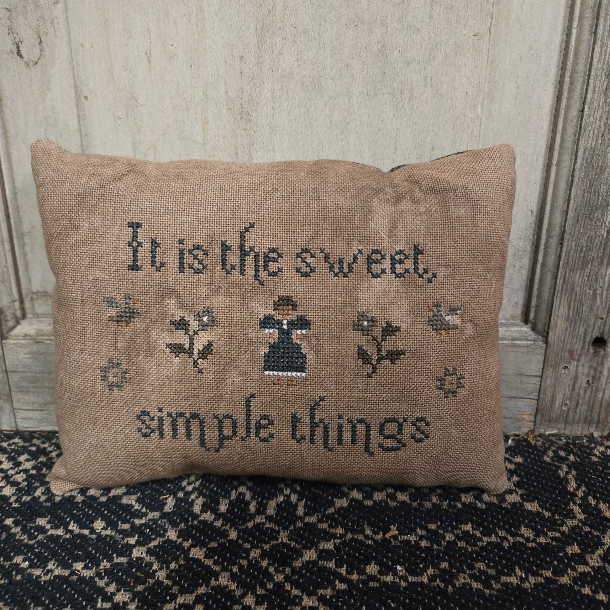 A rectangular cross stitch pillow with a floral design and the words 'It is the sweet simple things' embroidered on it, displayed on a textured surface.