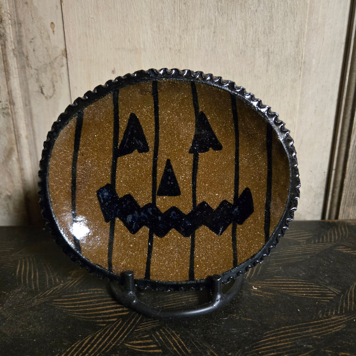A round black-rimmed decorative platewith a glittery gold interior featuring a black jack-o'-lantern face and vertical black stripes, displayed on a stand against a light wood background.