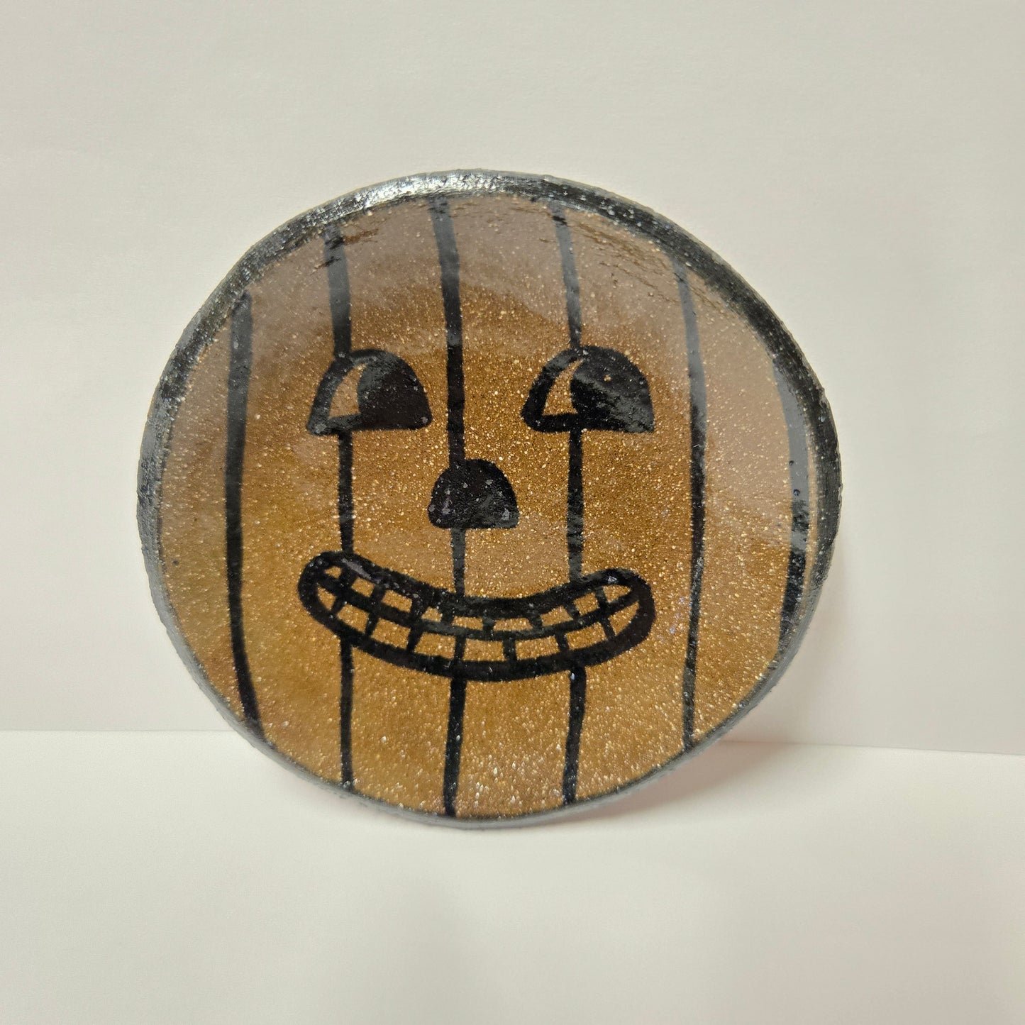 A round, hand-painted stoneware plate featuring a jack-o'-lantern face design on a glittery gold background with black vertical stripes and black facial features, set against a plain off-white background.