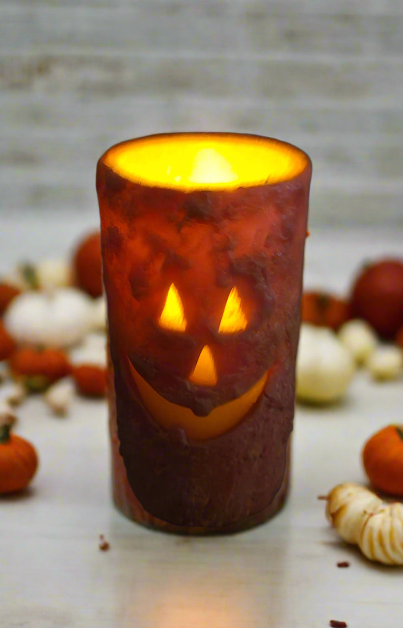 A burnt orange wax jack-o'-lantern votive candle with a carved face, lit and glowing, placed on a textured surface.