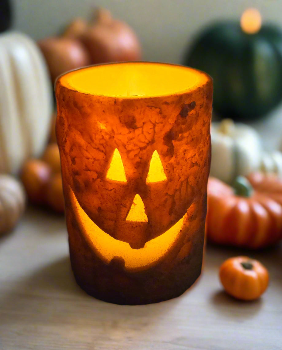 A battery-operated orange pillar candle with a carved jack-o'-lantern face that is glowing.