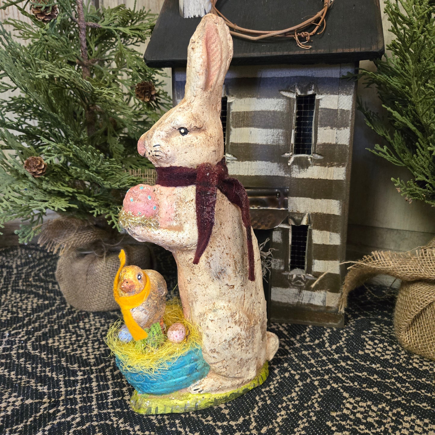 A handcrafted chalkware Easter decoration featuring a bunny with a chick in a basket, with a striped background.