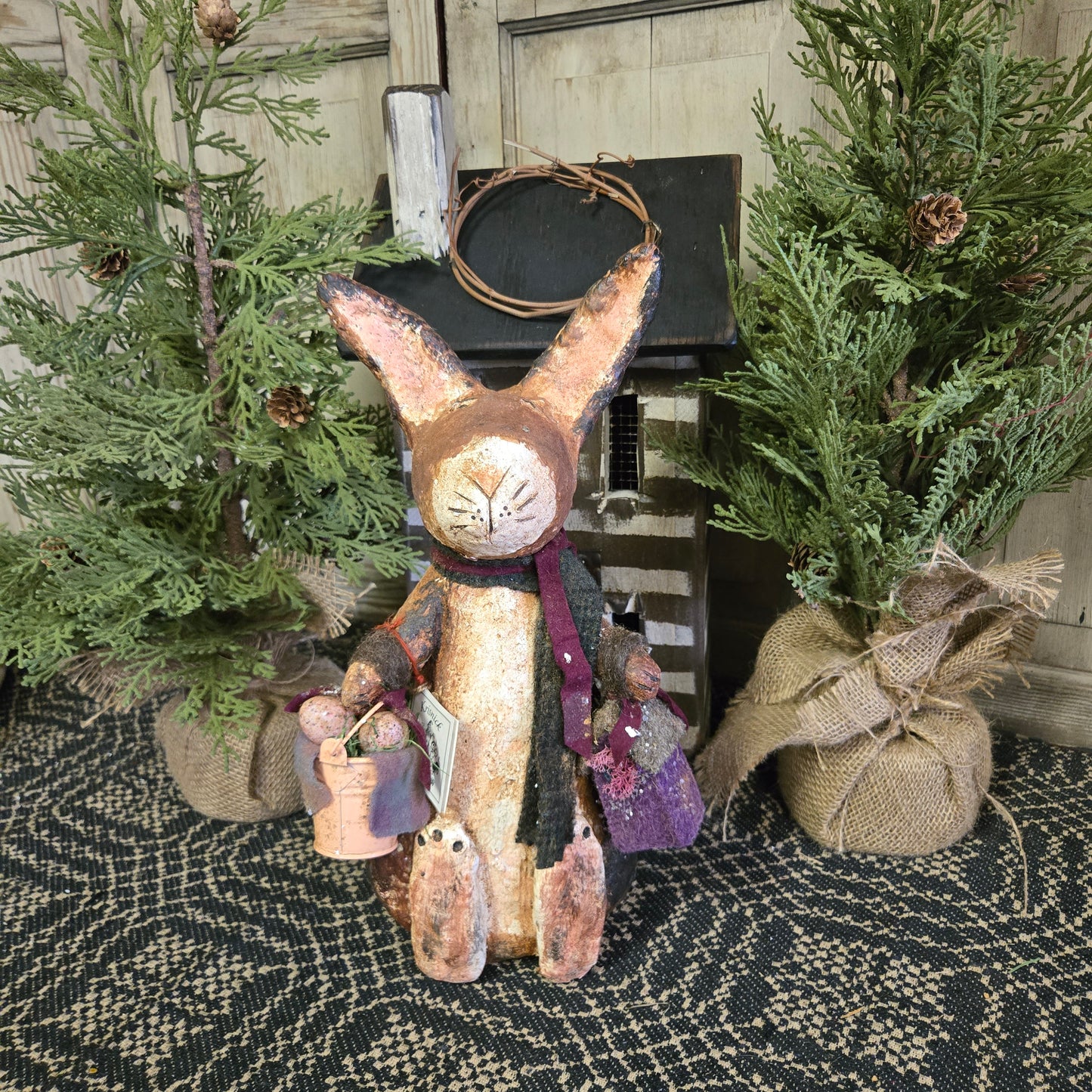 A handcrafted figurine of a Krisnick bunny holding a pail of eggs, positioned among decorative greenery.