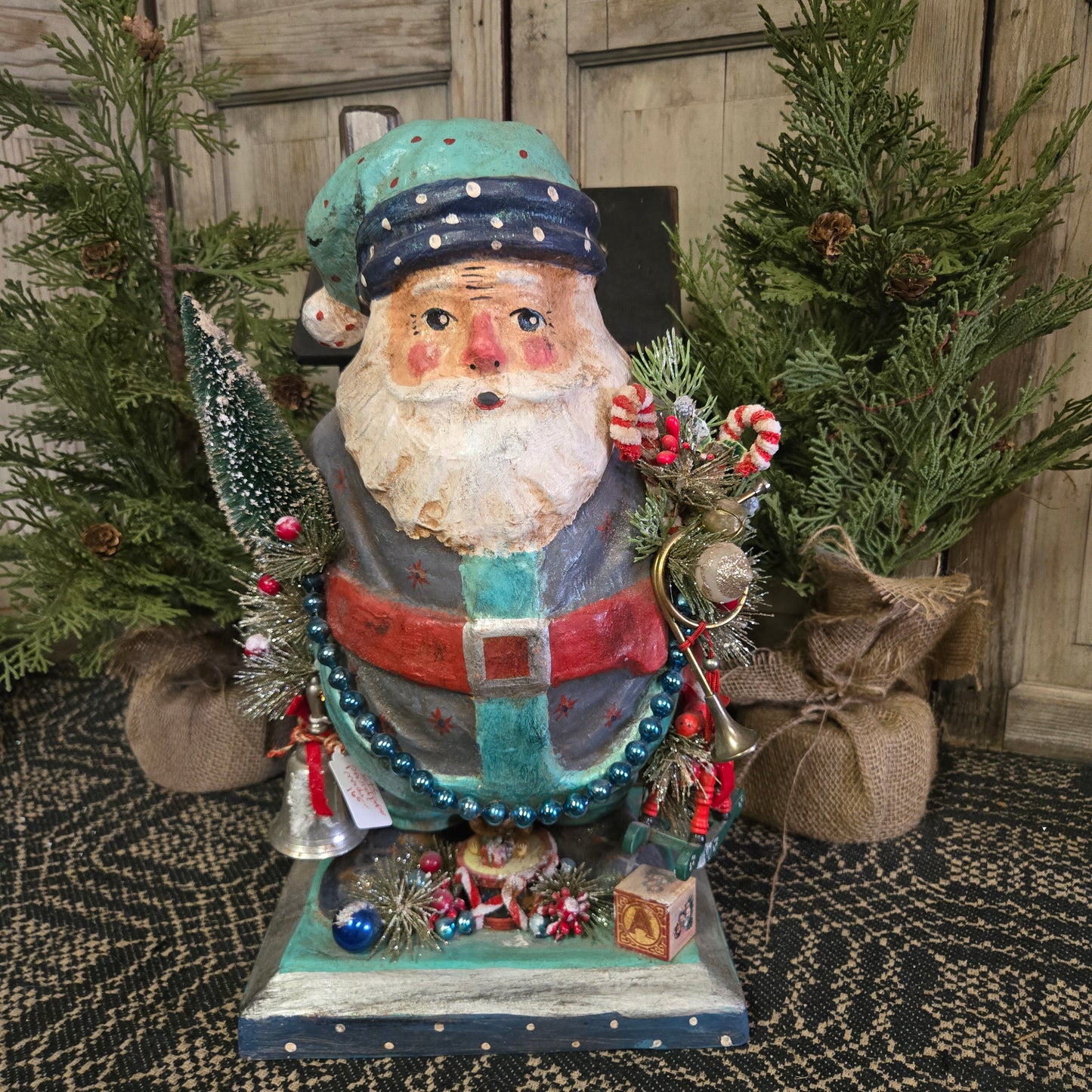 A handcrafted paper mache Santa figurine with a teal hat and toys, displayed against a rustic backdrop.