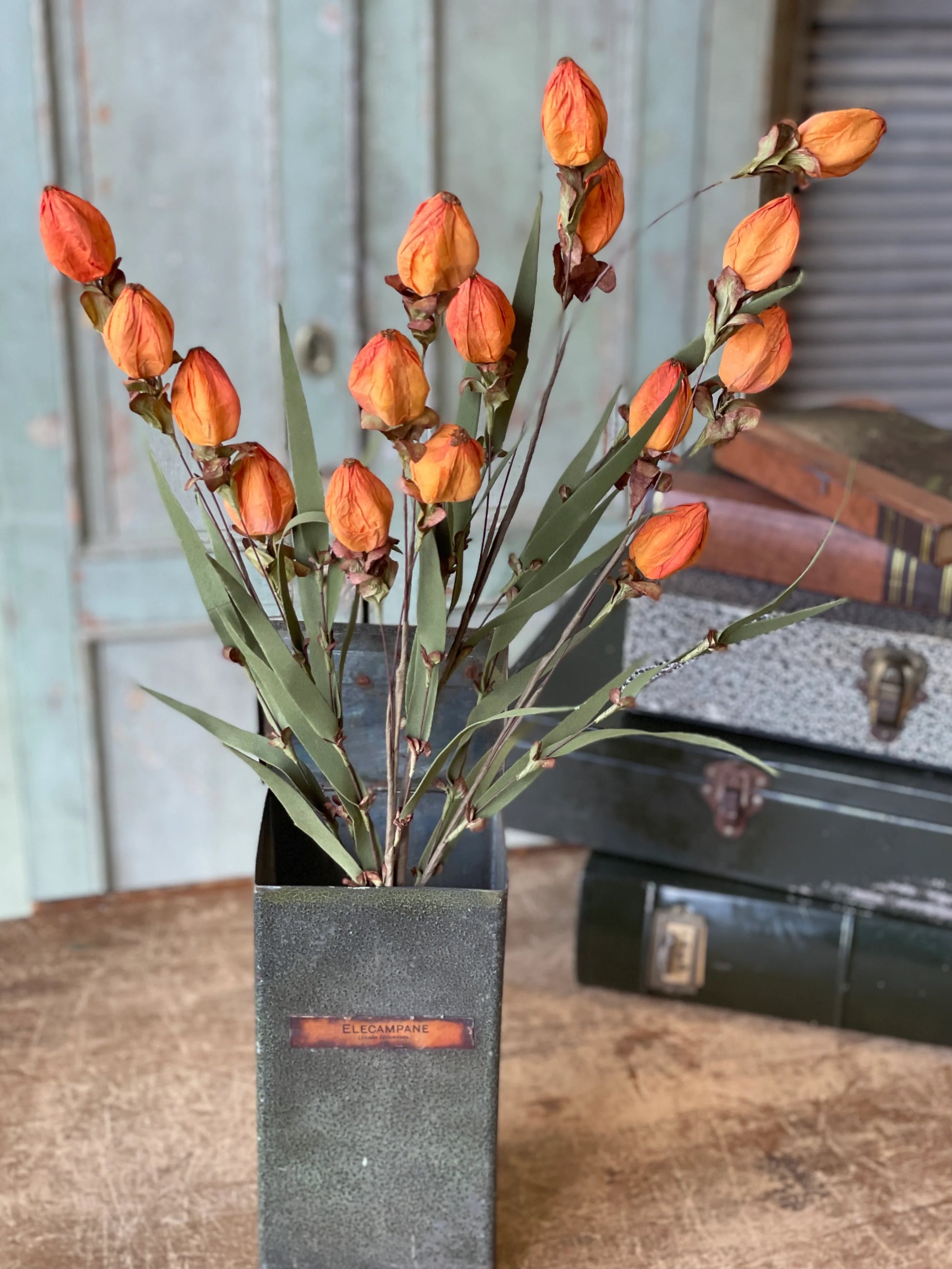 A vase containing a spray of orange lantern-like artificial buds nestled into greenery, placed on a rustic wooden surface.