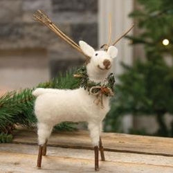 A felted white standing reindeer ornament with twig legs and a greenery wreath, displaying a smiling face and rustic twig antlers, placed on a wooden surface with a blurred Christmas tree in the background.