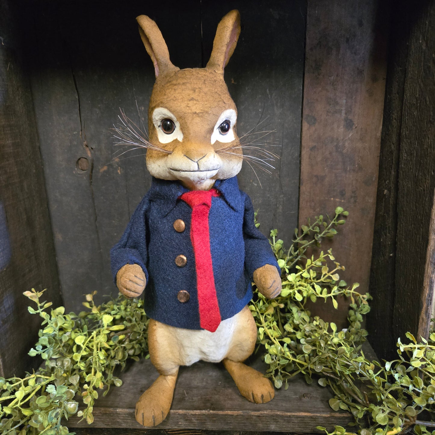 Stuffed animal resembling a rabbit in a blue outfit with a red collar on a wooden surface with greenery.