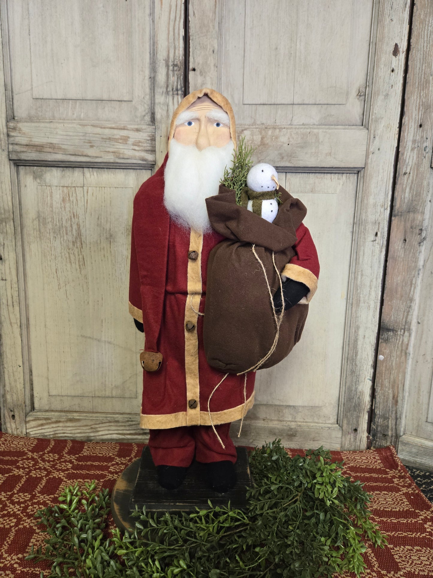 A large primitive Santa figurine wearing a red coat and holding a burlap sack with a snowman inside. The figurine is placed on a platform with holiday-themed decoration around it.