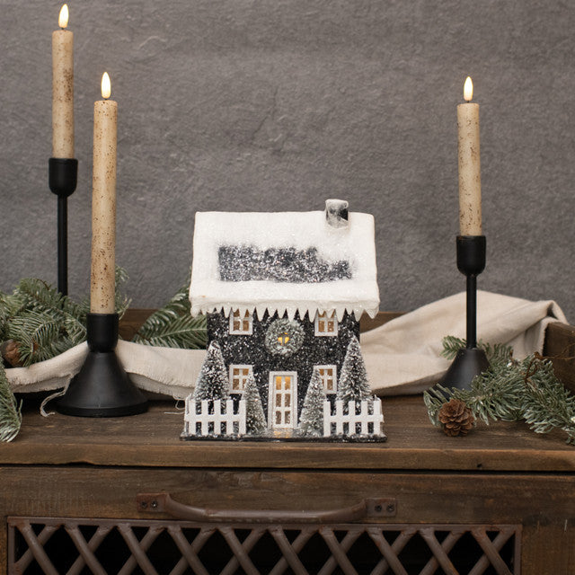 A decorative lighted black colonial home with a snowy roof, placed on a wooden surface with candles on either side, surrounded by holiday decorations.