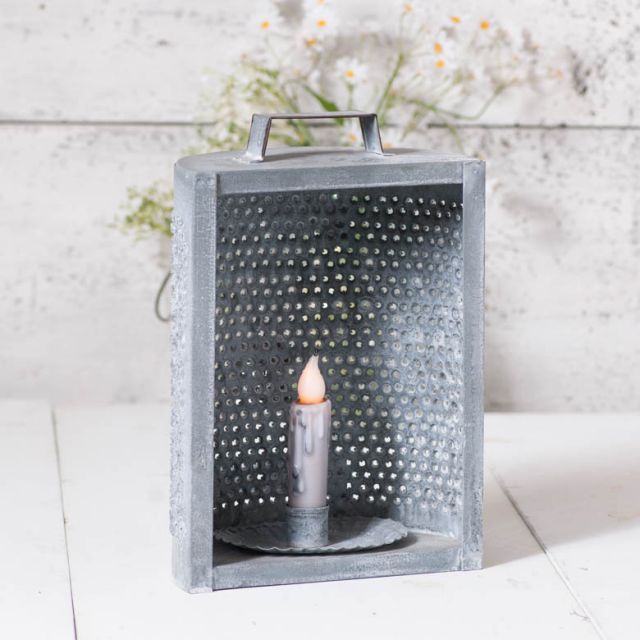 A weathered zinc candle holder with a grid design and a lit candle inside, placed on a white surface with a vintage background.