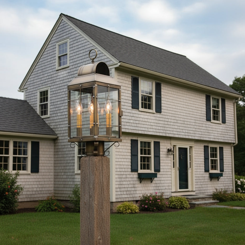 A weathered brass post lantern with clear glass and three internal light bulbs mounted on post in front of salt box style home.