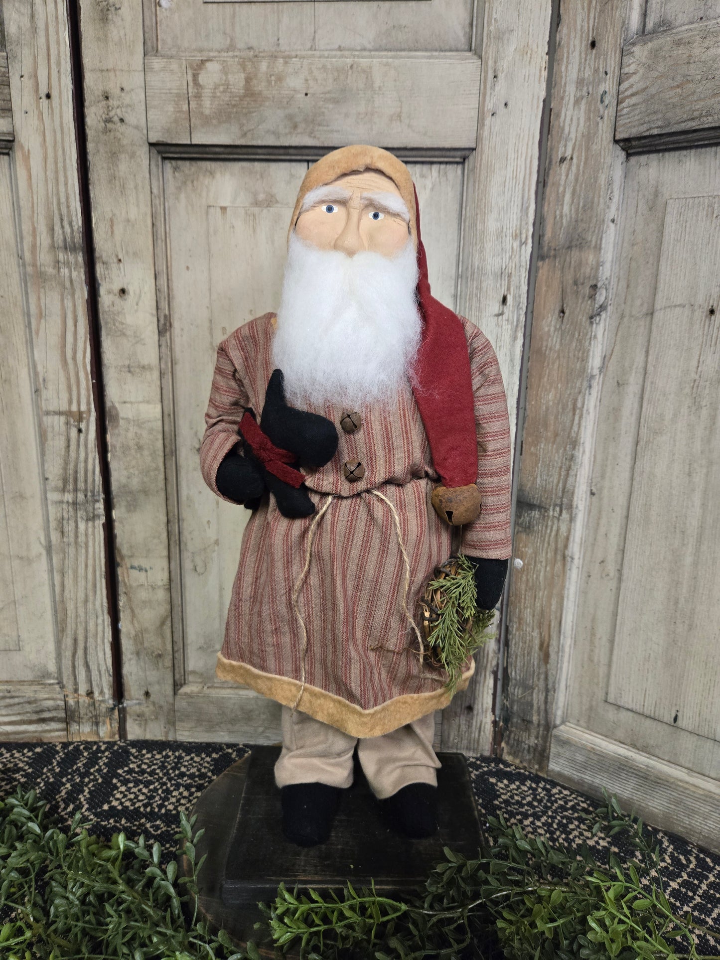 A medium-sized, primitive-style Santa figurine wearing a red and black striped coat and black hat, holding a dog on a leash. The figurine is standing against a wooden backdrop.