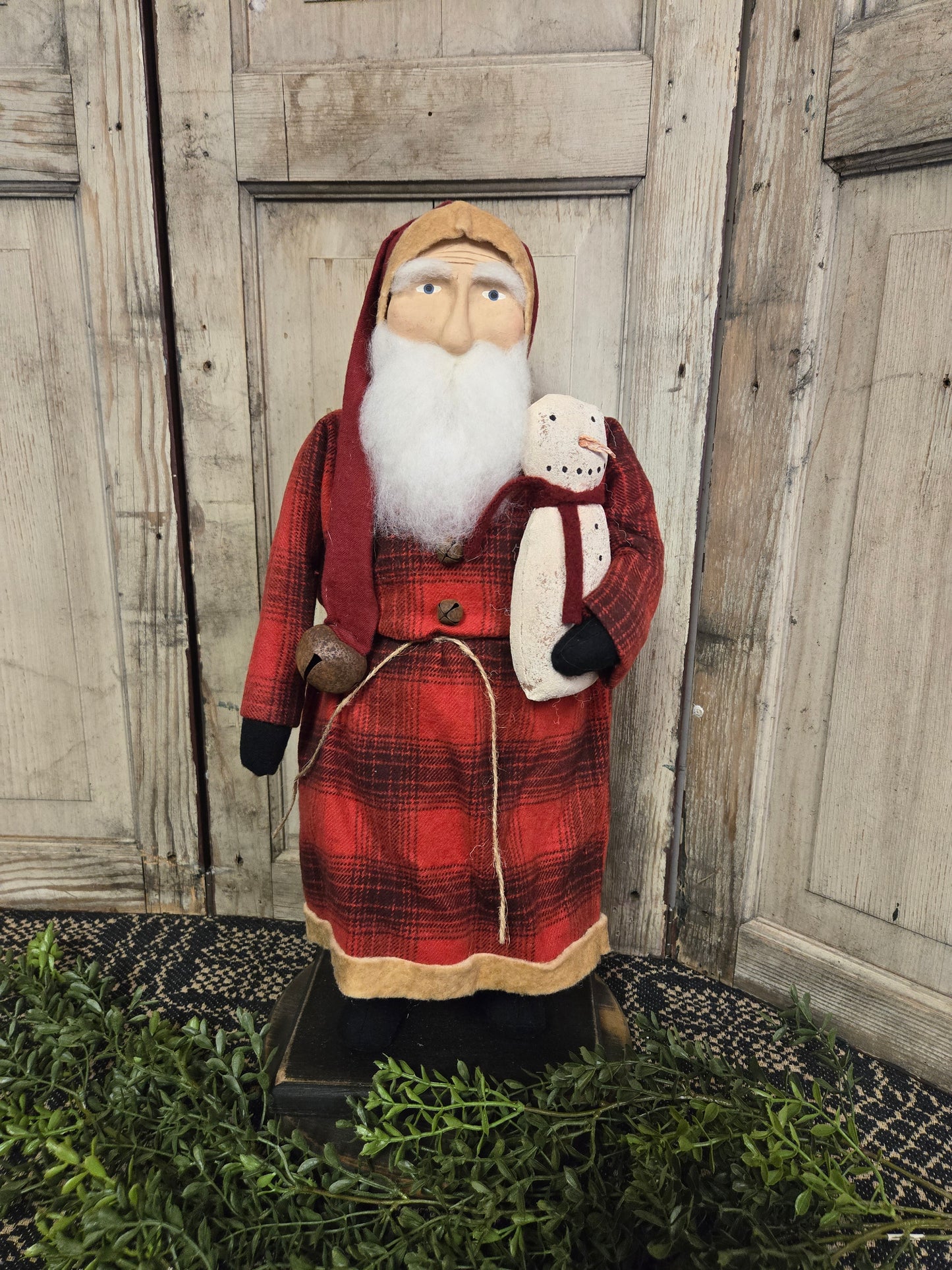 A medium-sized, handcrafted wooden figurine of Santa Claus wearing a red plaid coat, holding a snowman, with a backdrop of a wooden door and greenery.