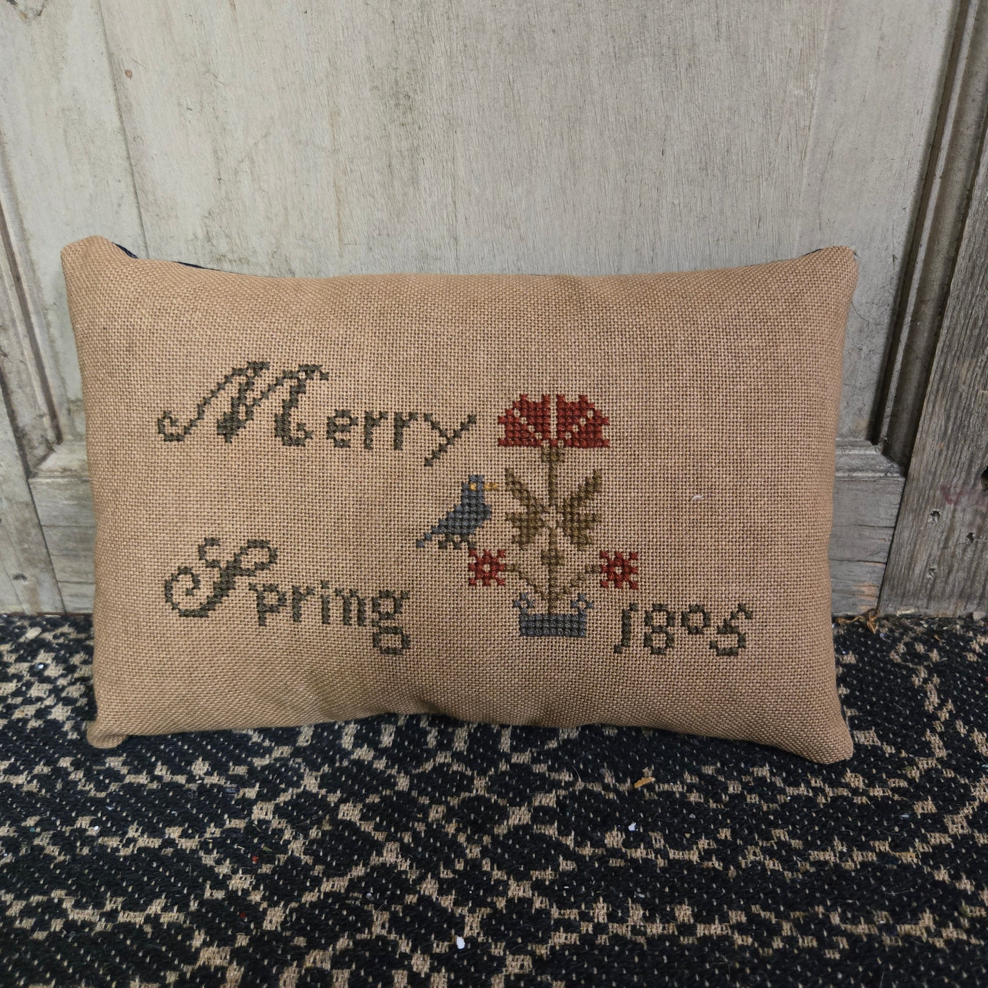 A rectangular cross stitch pillow with a floral design and the year '1805' embroidered on it, displayed on a textured surface.