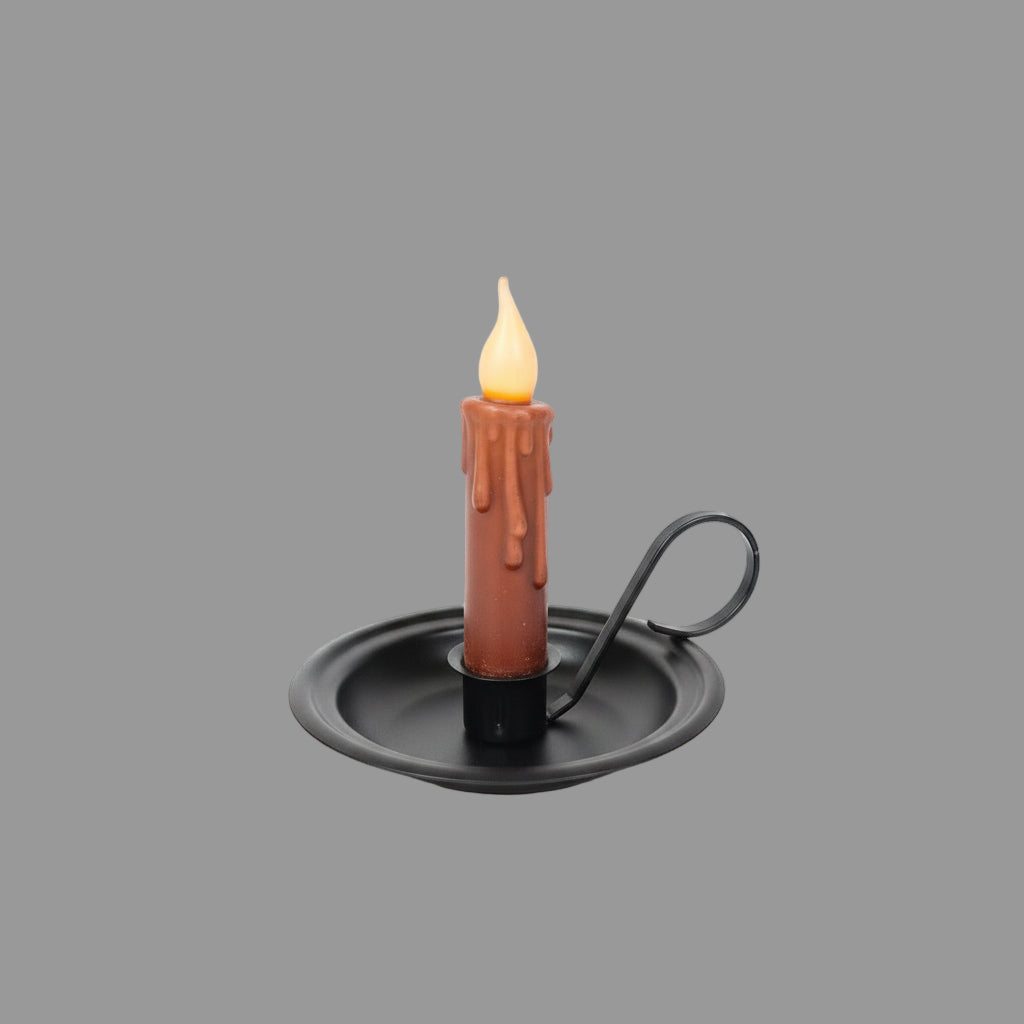 A metal taper candle holder with a lit candle surrounded by green foliage on a round table.