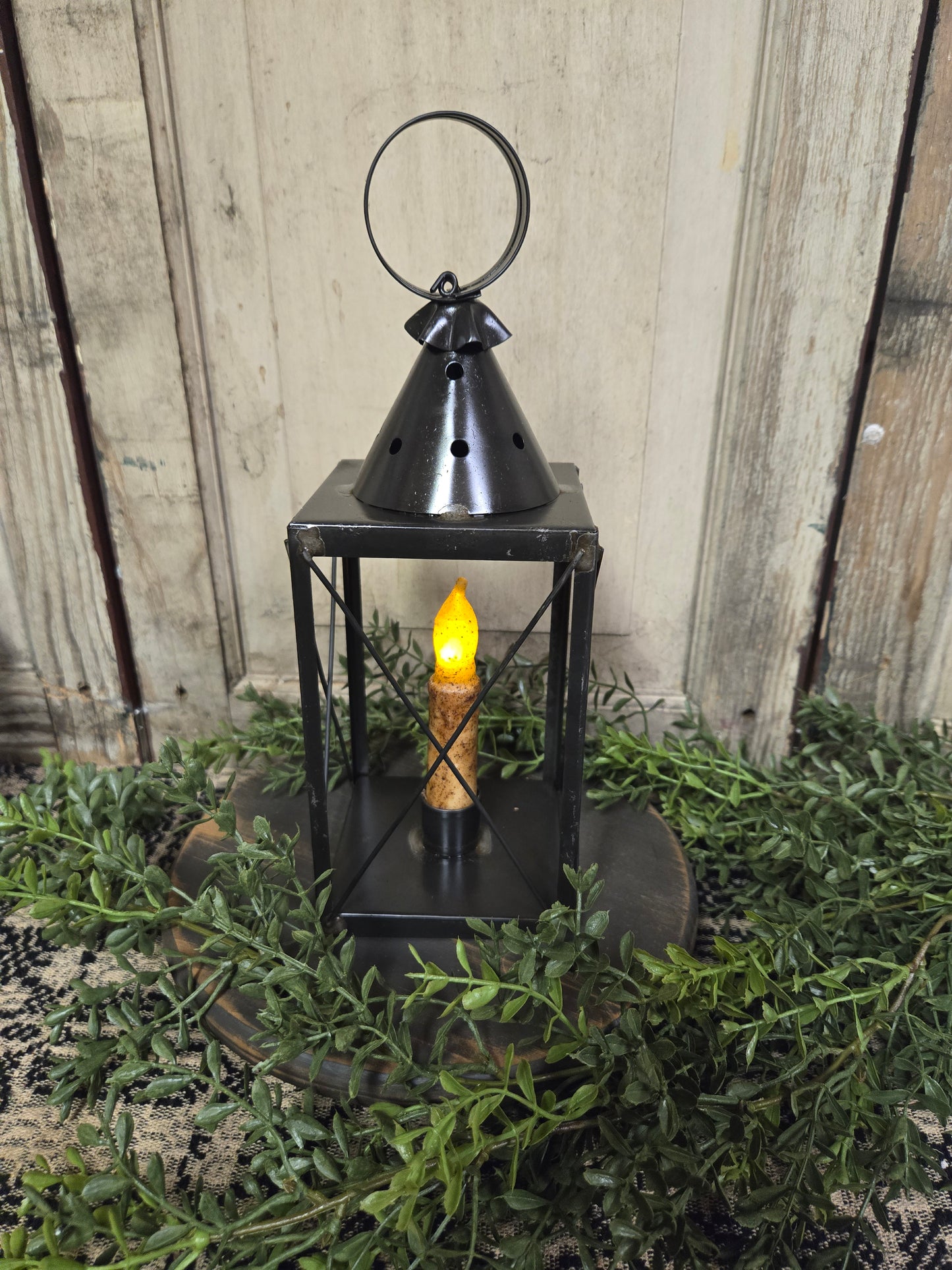 A black mini tin lantern with cross wires, displaying a lit candle inside, set on a bed of greenery.