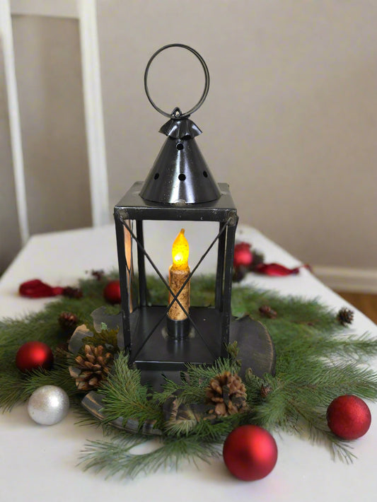 A black mini tin lantern with cross wires, displaying a lit candle inside, set on a bed of greenery.