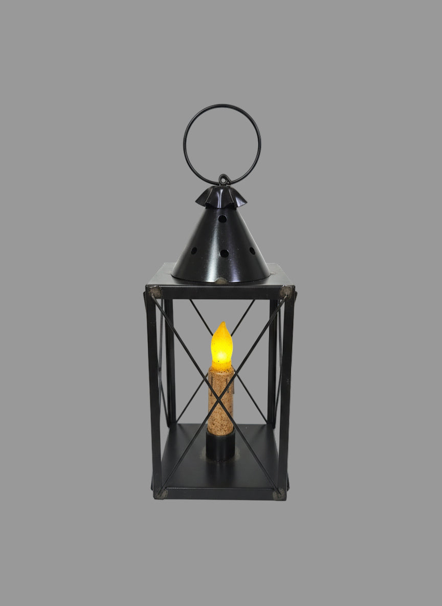A black mini tin lantern with cross wires, displaying a lit candle inside, set on a bed of greenery.