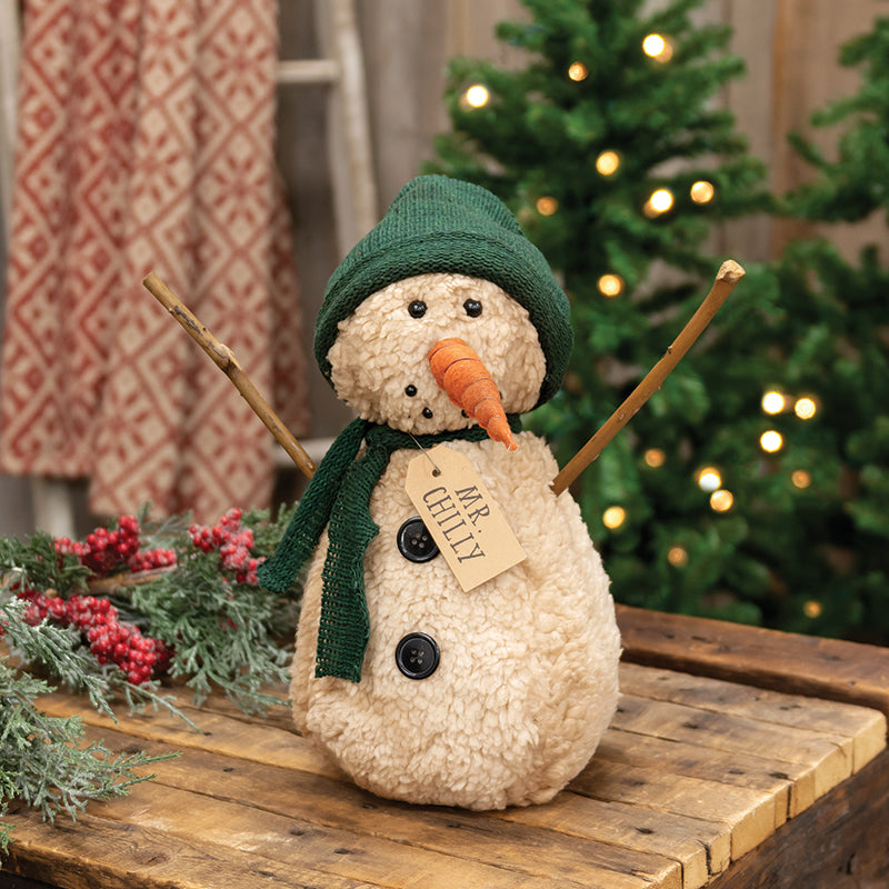 A rustic stuffed snowman doll with a dark green scarf and hat, an orange carrot nose, and the name 'Mr. Chilly' on a brown paper tag.