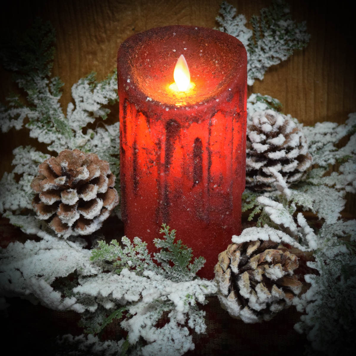 A red pillar candle with a moving flame effect, surrounded by faux snow and pine cones.