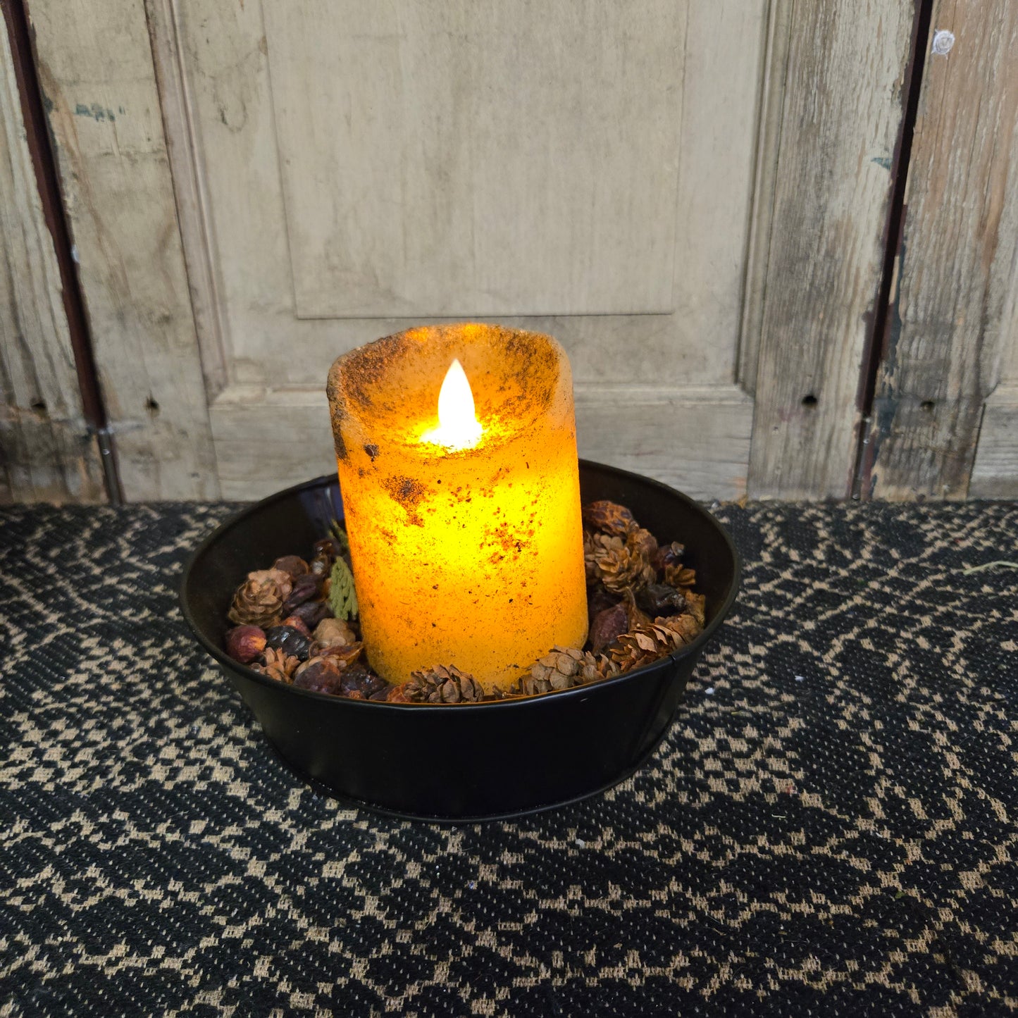 An LED pillar candle with a moving flame effect, placed on a black dish with a lit texture, set against a wooden background.