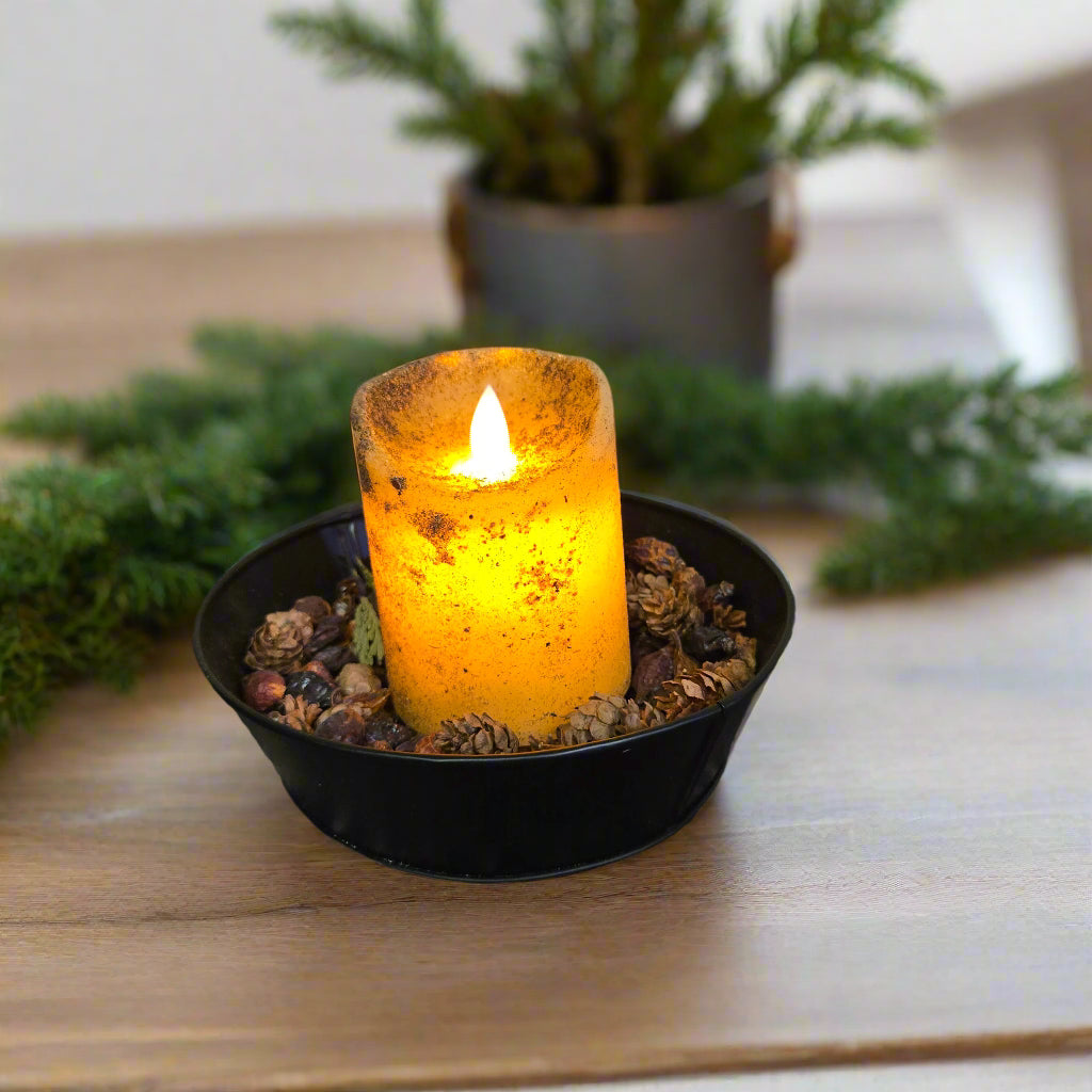 An LED pillar candle with a moving flame effect, placed on a black dish with a lit texture, set against a wooden background.