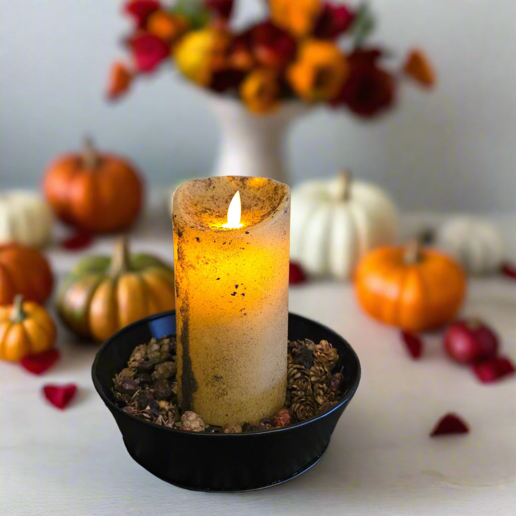 A moving flame pillar candle with a beige color and a rustic texture, placed in a black holder with a base of pebbles, set against a wooden background.