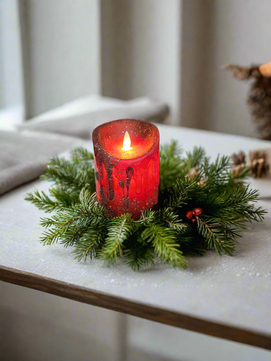 A red mica pillar candle with an illuminated flame, placed on a round, dark wicker mat, with a rustic wooden background and a door in the background.