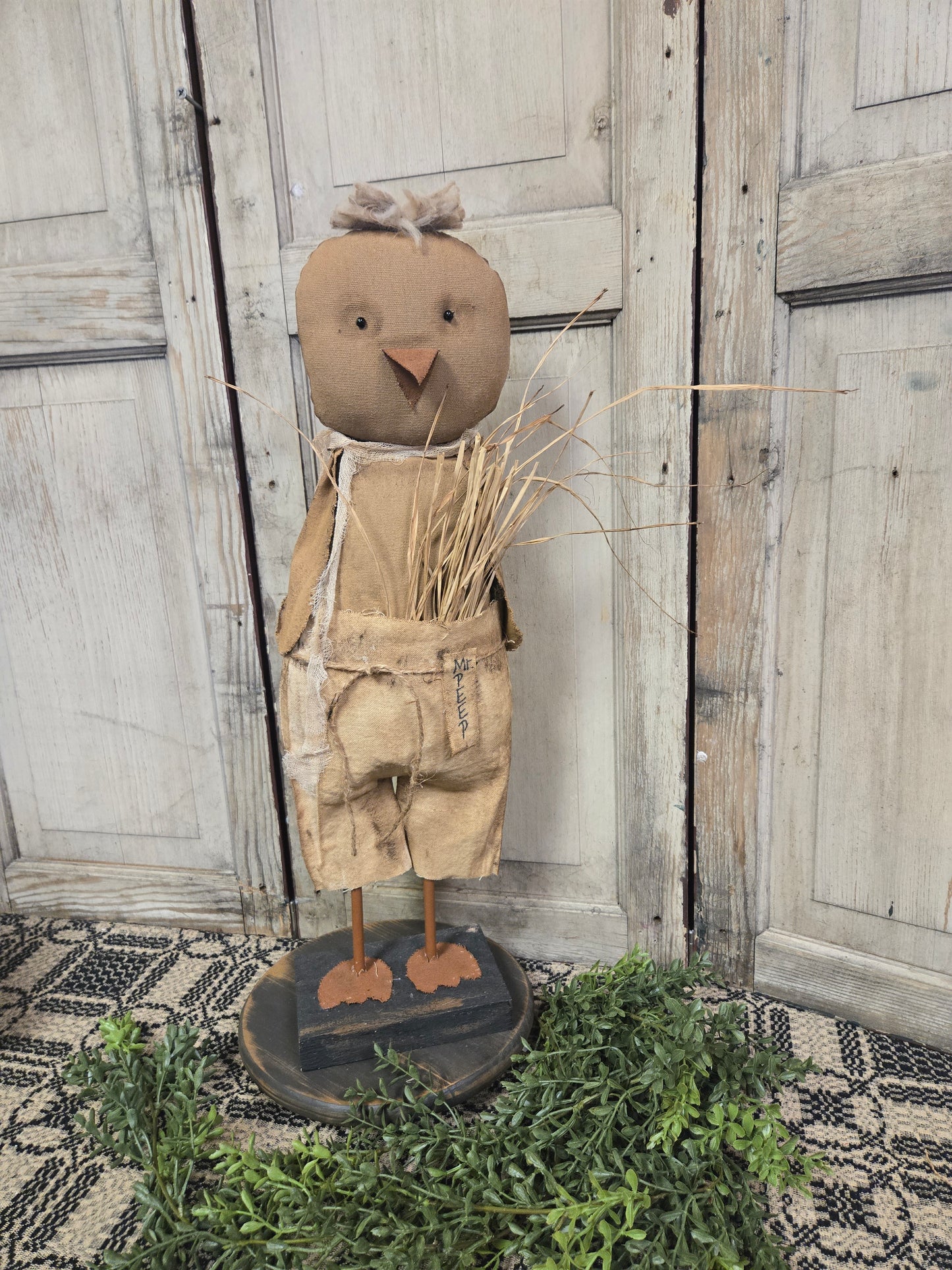 A sculpture of a chicken with a human-like face, wearing overalls and holding a straw hat, standing on a base decorated with greenery.