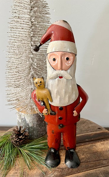 Wooden Santa Claus figurine holding a cat on a rustic wooden surface with a textured wall background.
