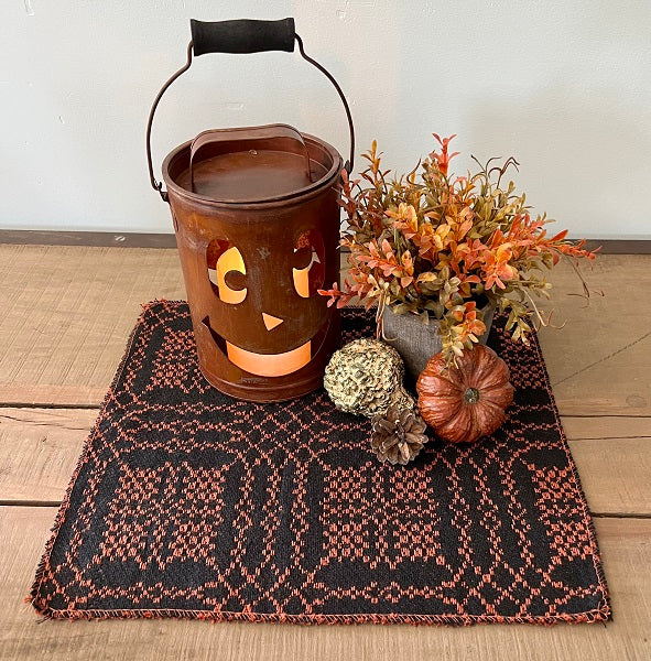 A square placemat with a black and rust color scheme, featuring a jack-o-lantern design, placed on a wooden surface. Autumn-themed decorations, including pumpkins and dry leaves, are placed on top of the placemat.