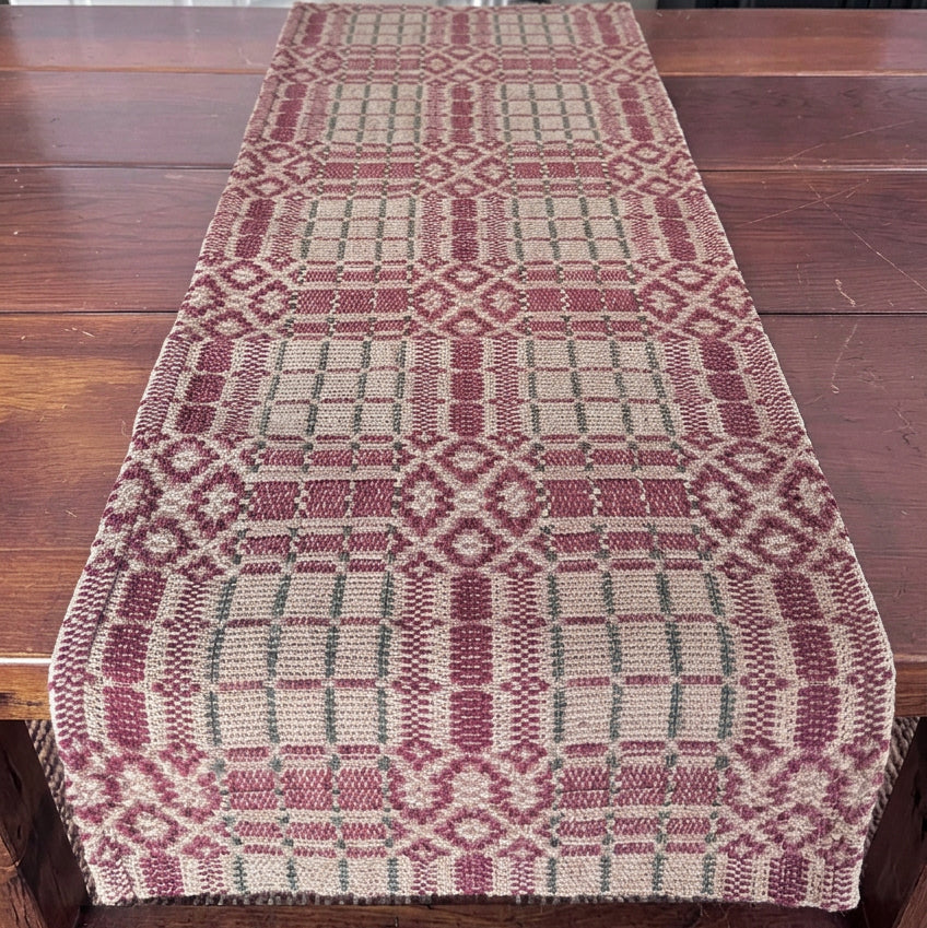 A woven table runner with a geometric pattern in burgundy, beige, and green, styled on a dark wooden table with Christmas raven figurines and a candle.