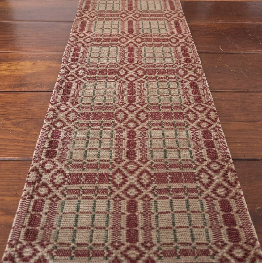 A woven table runner with a geometric pattern in burgundy, beige, and green, styled on a dark wooden table with Christmas raven figurines and a candle.