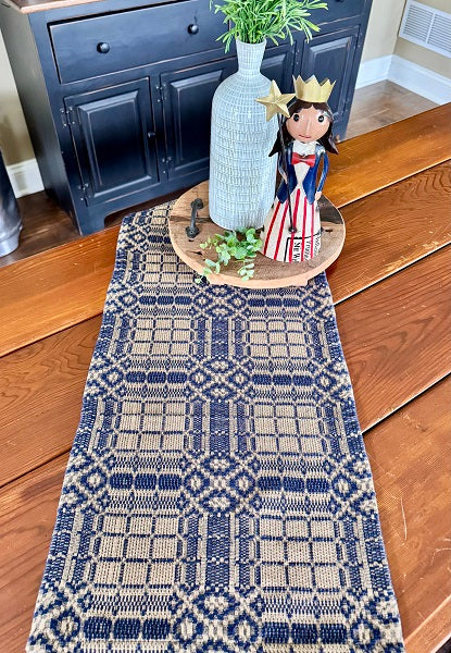Decorative table runner with geometric pattern on a wooden floor, with a vase and figurine in the background.